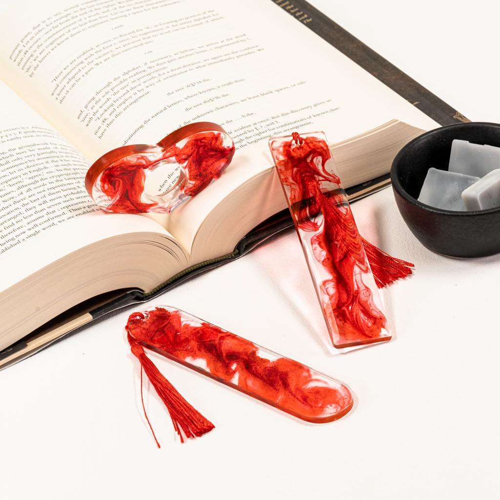 Red and clear glitter heart-shaped page holder propped on open book, along side an oval and rectangle bookmark with blood-like swirls, next to a dish of grey wax melts.