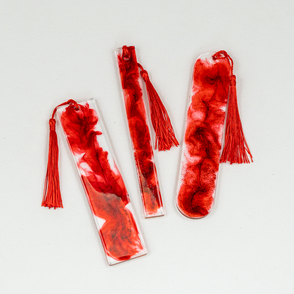 Three clear resin and red wisp bookmarks with glitter and red tassels on white background.
