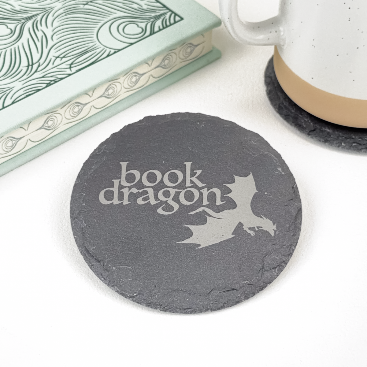 A round dark gray slate coaster with 'Book Dragon' and a dragon silhouette printed on it, styled on a white surface next to a mug and a book.