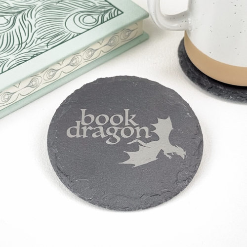 A round dark gray slate coaster with 'Book Dragon' and a dragon silhouette printed on it, styled on a white surface next to a mug and a book.