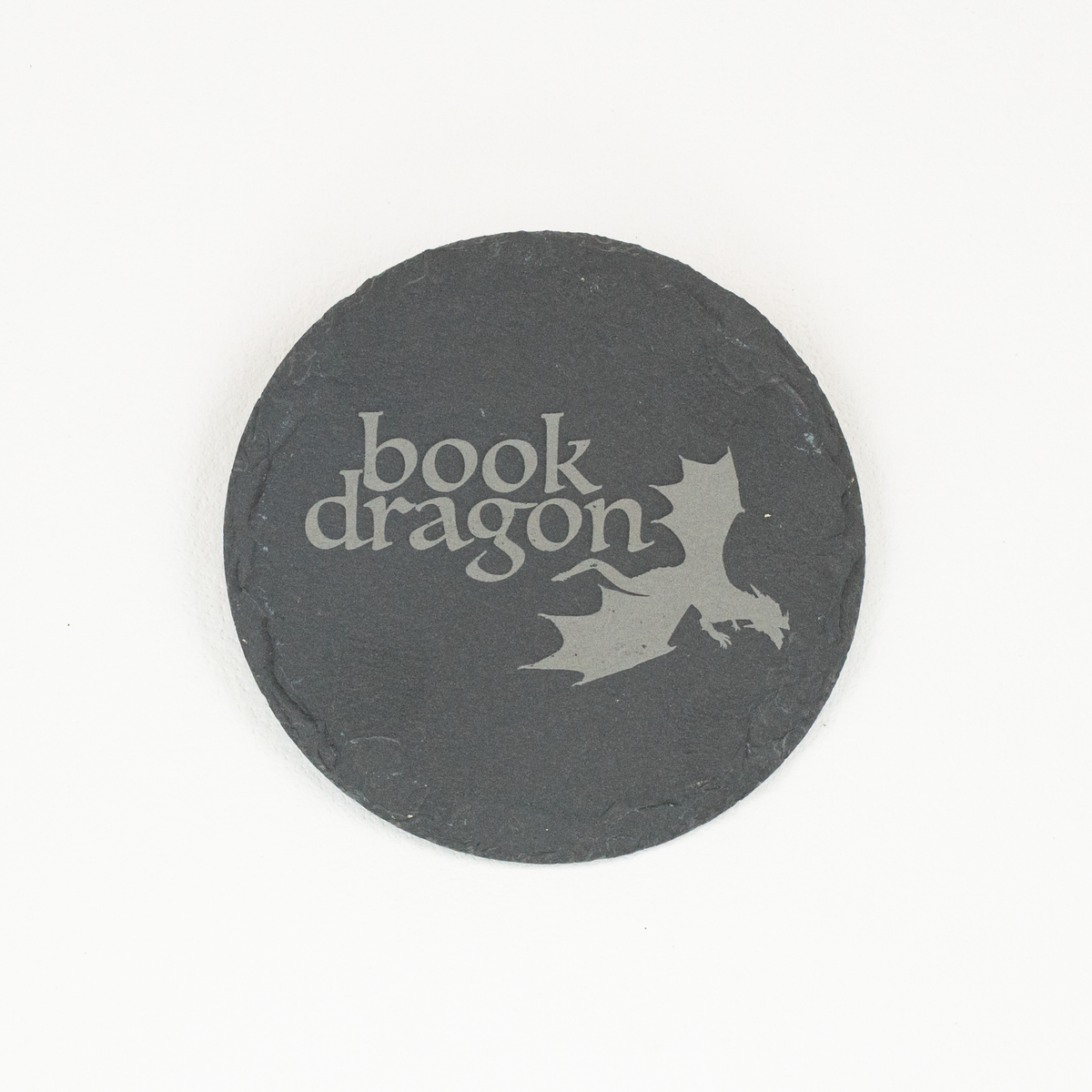 A round black slate coaster featuring the words 'Book Dragon' in a stylized light gray font and a matching dragon silhouette, shown on a plain white background.