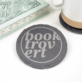 A round dark gray slate coaster with the words 'Booktrovert' in light gray lettering, styled on a white surface next to a patterned book and a white ceramic mug.