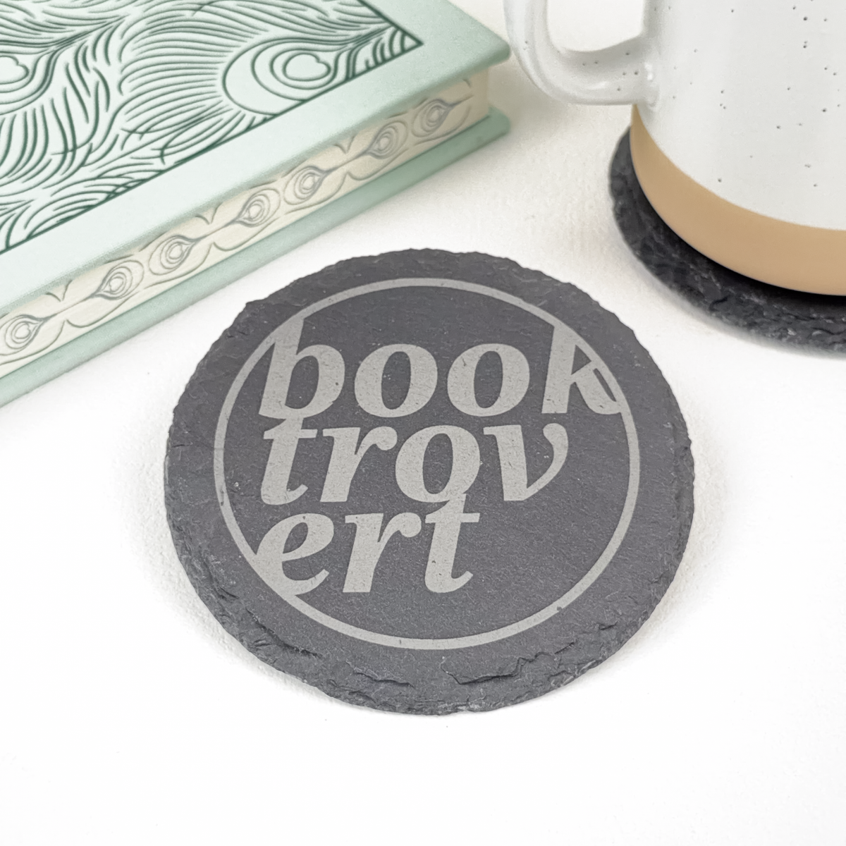A round dark gray slate coaster with the words 'Booktrovert' in light gray lettering, styled on a white surface next to a patterned book and a white ceramic mug.