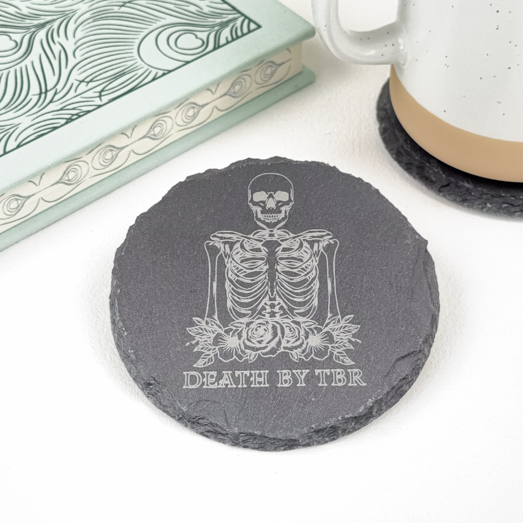 A round dark gray slate coaster featuring an engraved skeleton with roses and the text 'DEATH BY TBR', styled on a white surface with a book and a ceramic mug.
