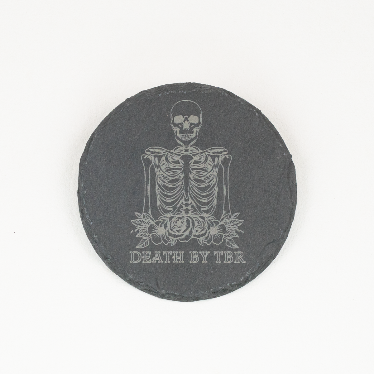 A round black slate coaster featuring a white engraved illustration of a skeleton with roses and the text 'DEATH BY TBR', shown on a plain white background.