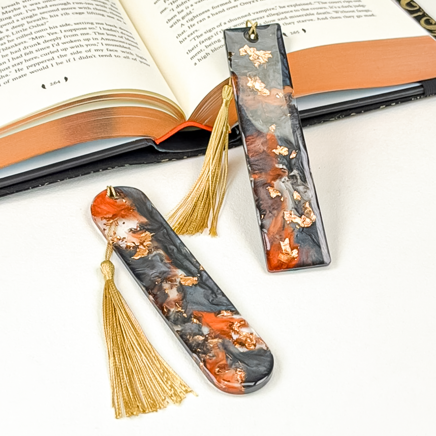 Two decorative resin bookmarks with gold tassels, featuring a marbled pattern of black, copper, and burnt orange, styled on an open book.