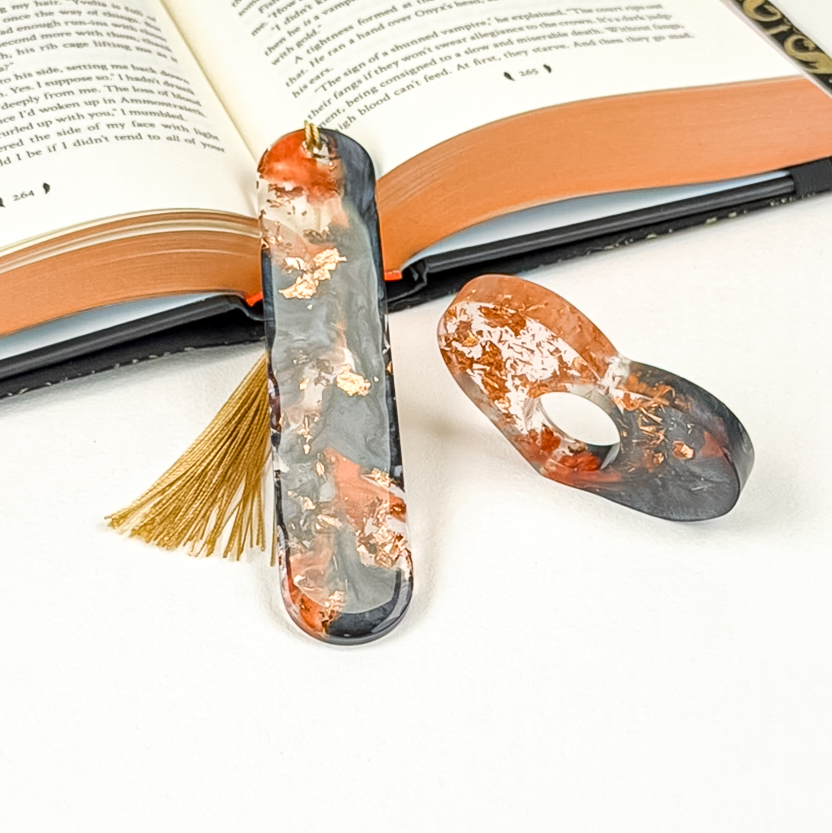 Two resin book accessories in a black, orange, and copper marbled pattern. One is an oval bookmark with a gold tassel, and the other is a heart-shaped thumb page holder placed on it's side with an open book on a white background.