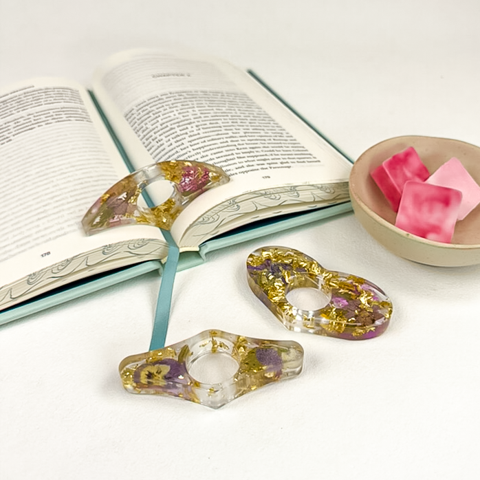 Three clear resin page holders with flowers and gold flakes in clear resin, styled next to open book and pink Amour wax melts.