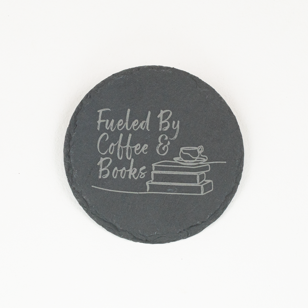 A round black slate coaster with white engraved text reading 'Fueled By Coffee & Books' and a small illustration of stacked books and a cup, shown on a plain white background.