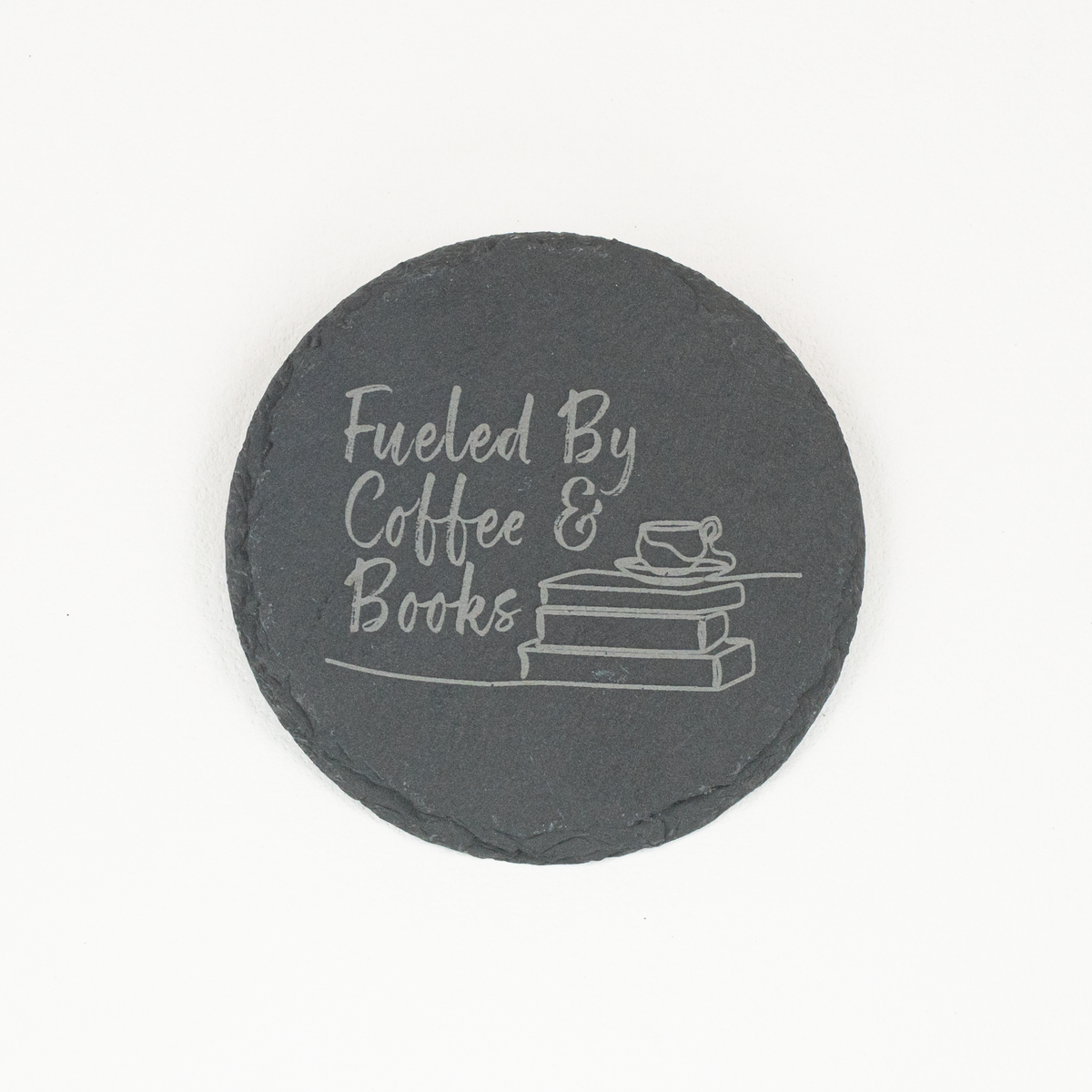 A round black slate coaster with white engraved text reading 'Fueled By Coffee & Books' and a small illustration of stacked books and a cup, shown on a plain white background.
