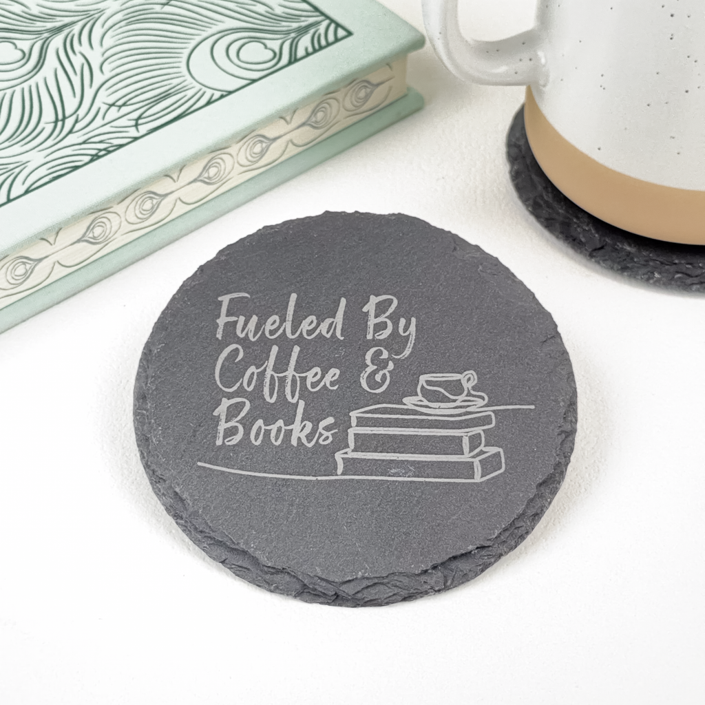 A round dark gray slate coaster engraved with the phrase 'Fueled By Coffee & Books' and a small book with a cup illustration, shown on a white surface with a book and mug partially visible in the background.