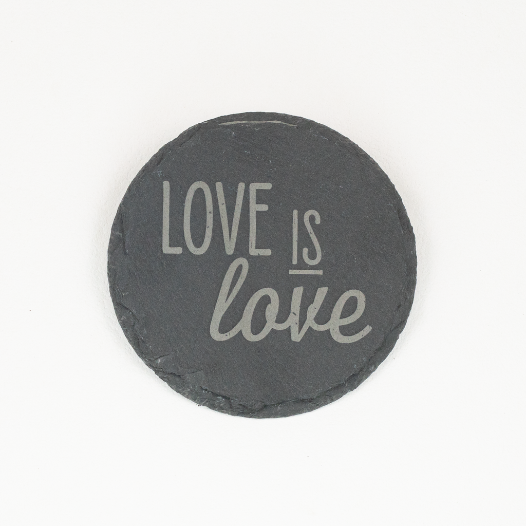 A round black slate coaster with the phrase 'LOVE IS love' printed in light gray lettering, shown on a plain white background.
