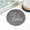 A round dark gray slate coaster with the words 'LOVE IS love' printed in light gray, shown on a white surface with a mug and a decorative book partially visible around it.