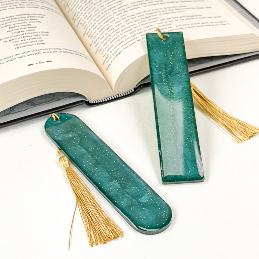 Two deep teal resin bookmarks with gold glitter and gold tassels propped against an open book.