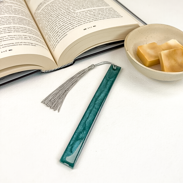 Single slim resin bookmark in deep teal with silver glitter and silver tassel, alongside open book and wax melts