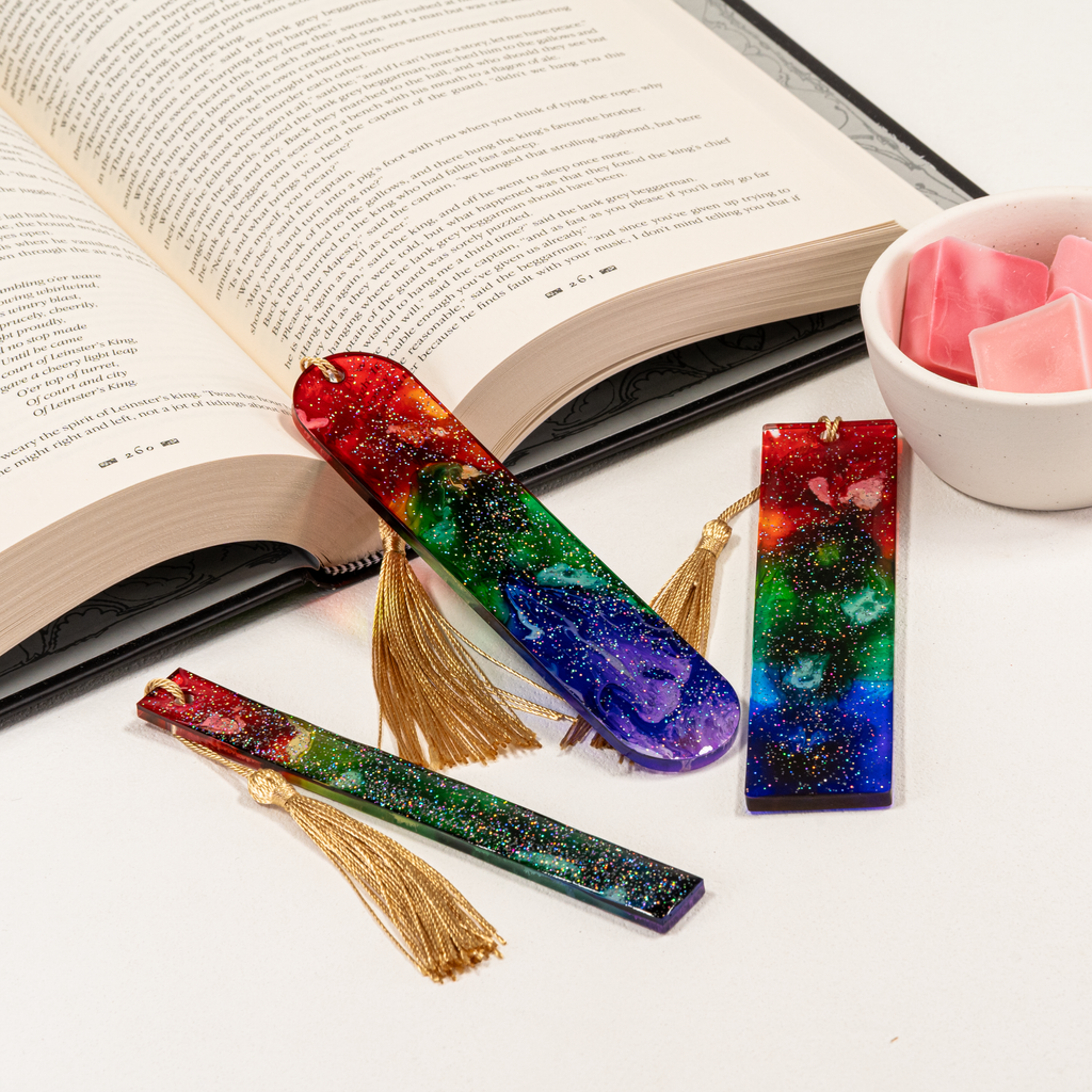 Three resin bookmarks in rainbow colors and glitter, arranged alongside an open book with a bowl of wax melts.