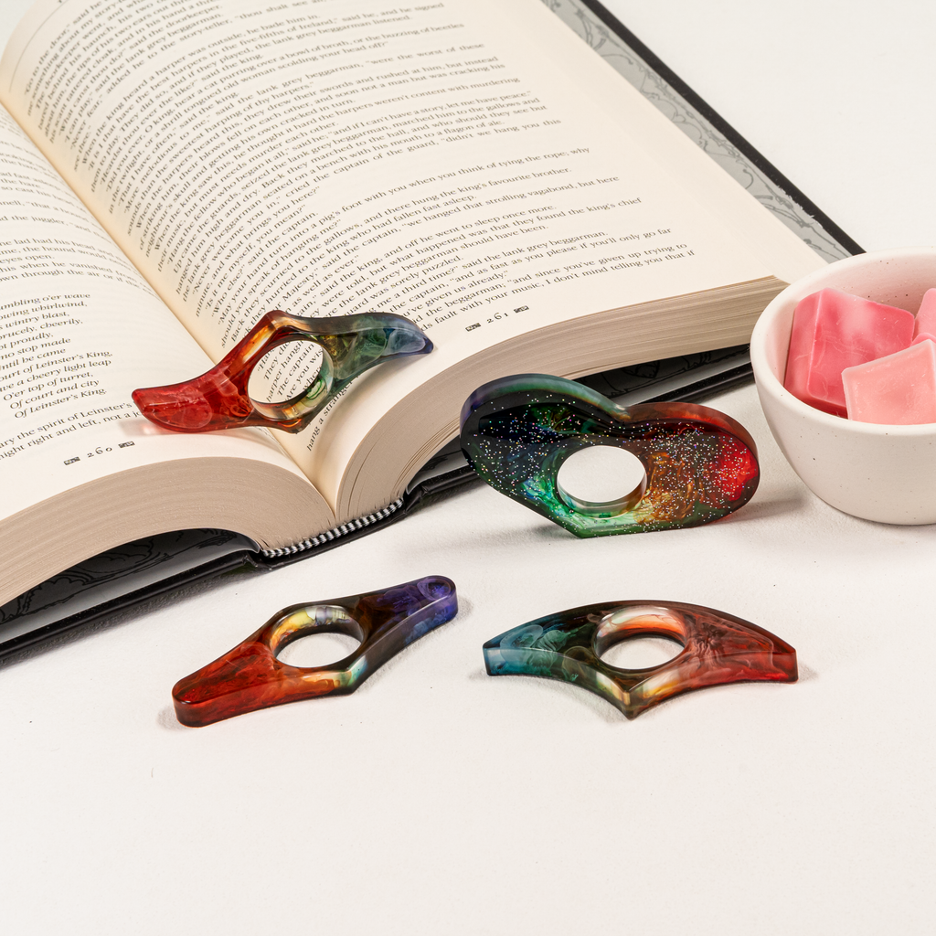 Four various shaped resin page holders with rainbow colors, glitter, and wisps of colors in translucent resin. Styled next to a book with a bowl of wax melts.
