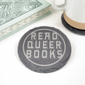 Single slate round coaster with "Read Queer Books" in stylized font engraved on surface. Styled next to a closed book and ceramic mug.