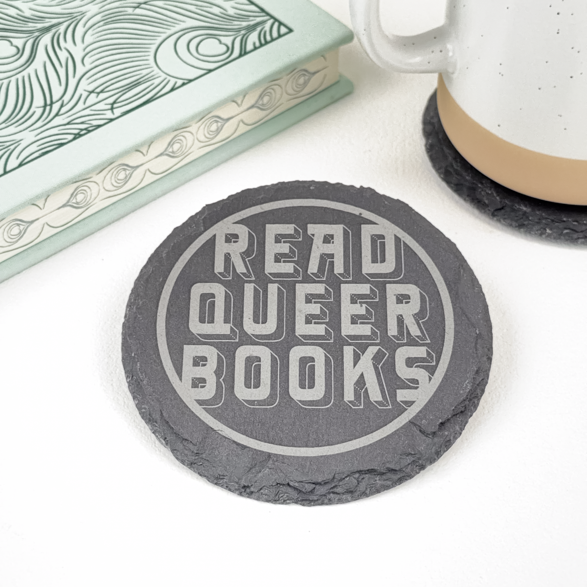 Single slate round coaster with "Read Queer Books" in stylized font engraved on surface. Styled next to a closed book and ceramic mug.