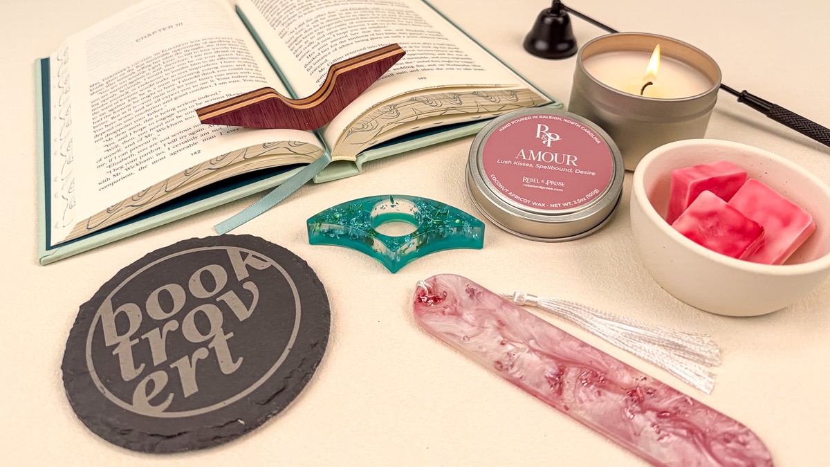 A styled flat-lay of book-themed self-care and reading accessories on a cream background: an open book with a wooden bookmark, a teal resin page holder, a pink tassel bookmark, a round slate coaster reading 'BOOKTROVERT', a pink candle tin labeled 'Amour', pink wax cubes in a white bowl, and a black candle extinquisher.