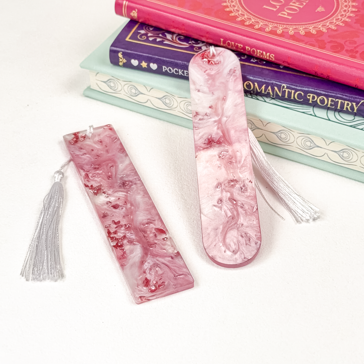 Two pink and white marbled resin bookmarks with white tassels, displayed on a white surface in front of a stack of decorative books.