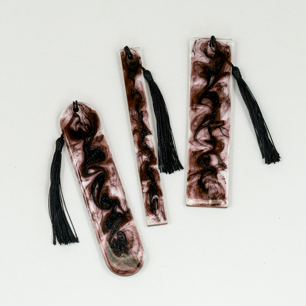 Three various shaped resin bookmarks with black tassels, featuring a swirling dark brown and black pattern, displayed on a clean white surface.