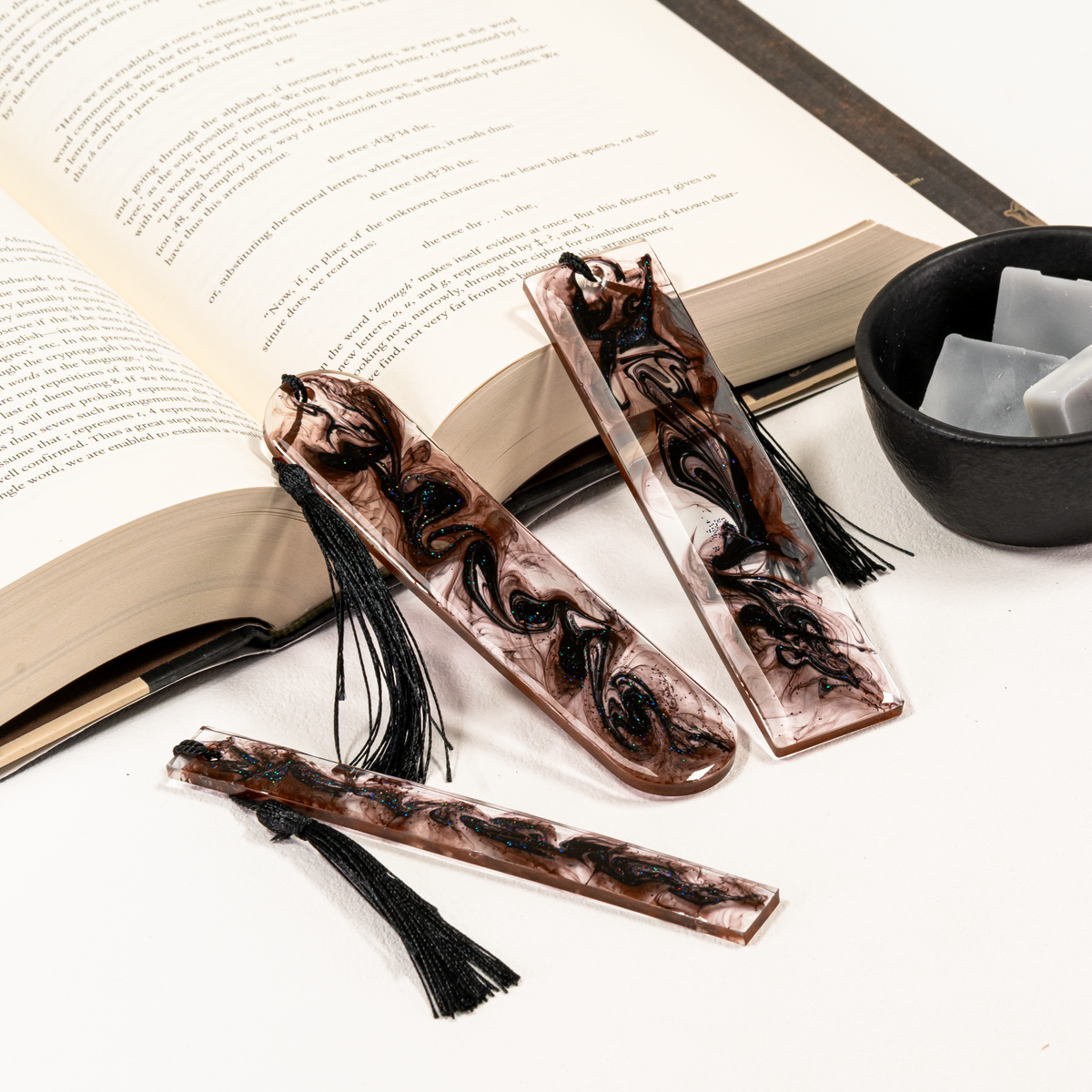 Three resin bookmarks with black tassels are arranged on an open book. The bookmarks have a swirling marbled pattern in translucent smoky gray tones.