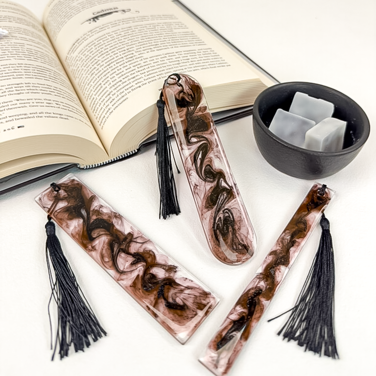 Set of three various sized clear resin bookmarks with swirls of black and glitter and black tassels. Styled next to an open book and bowl of wax melts.
