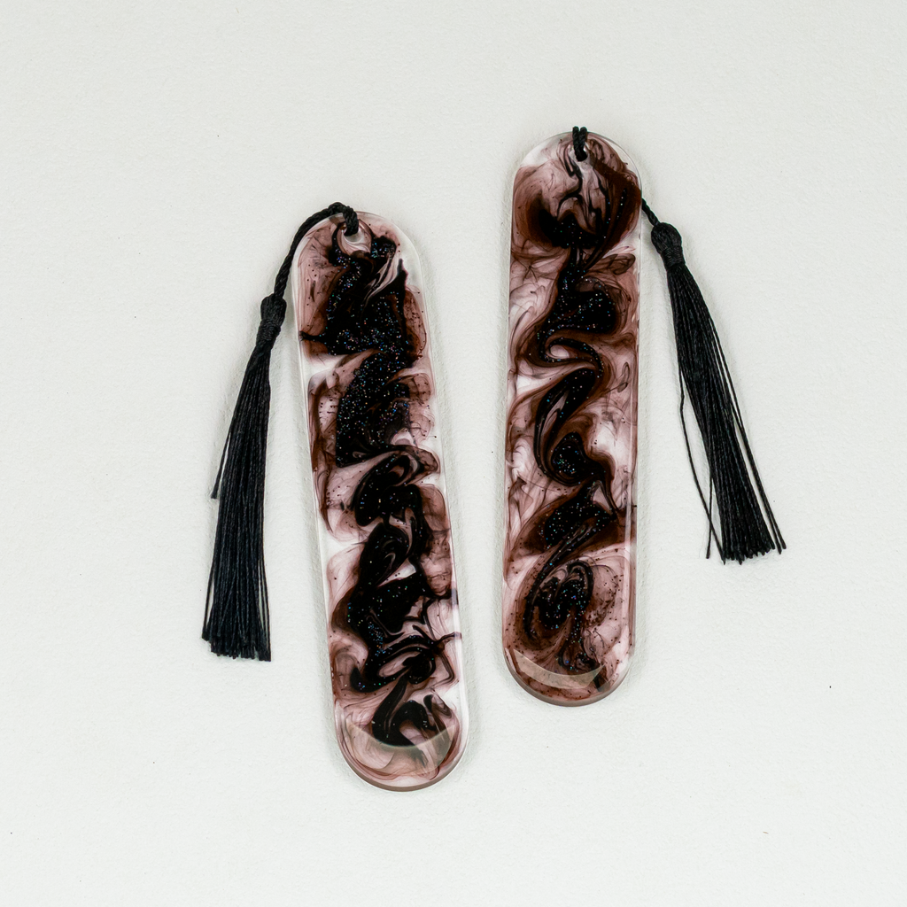 Two oval shaped resin bookmarks with black swirls and glitter, and black tassels.