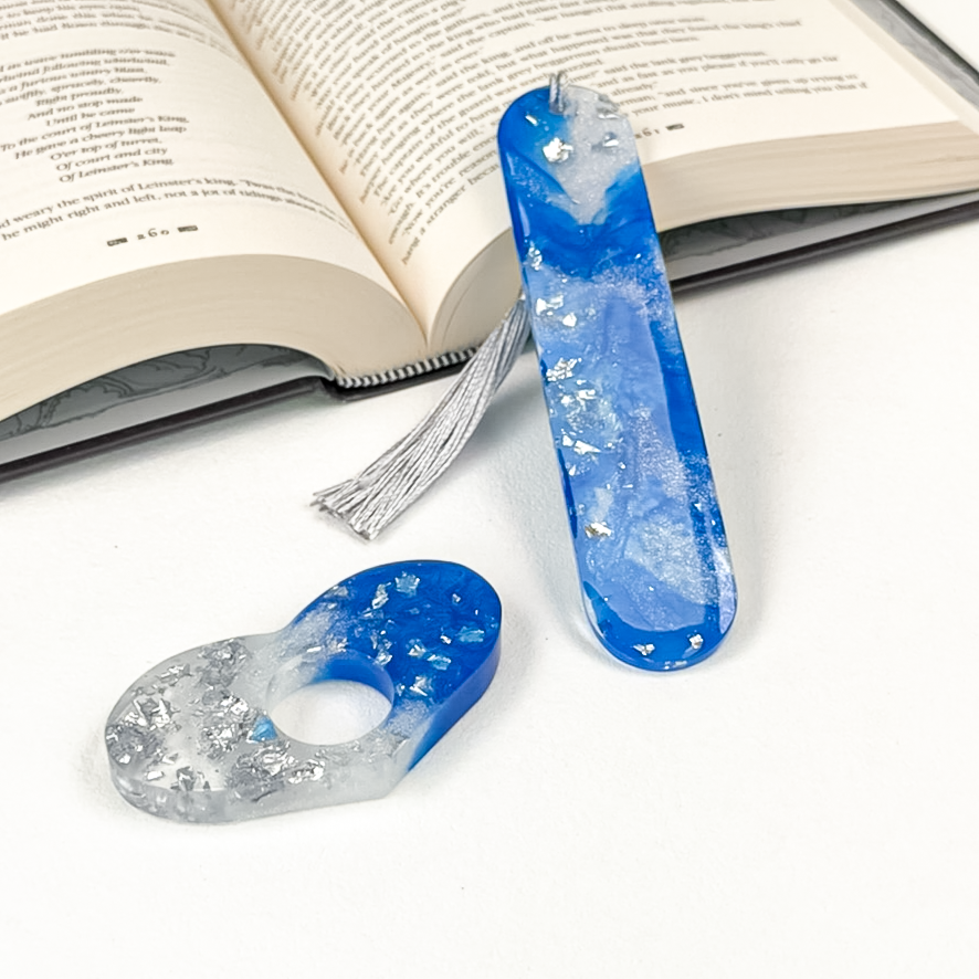 A heart-shaped resin page spreader and oval bookmark, both with blue and silver marbled pattern, displayed on white surface with an open book in the background. 