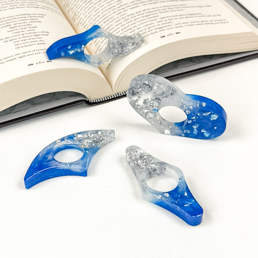 Four blue, white, and silver resin page spreaders shaped like a blade, a heart, a fan, and a wing, set on an open book.