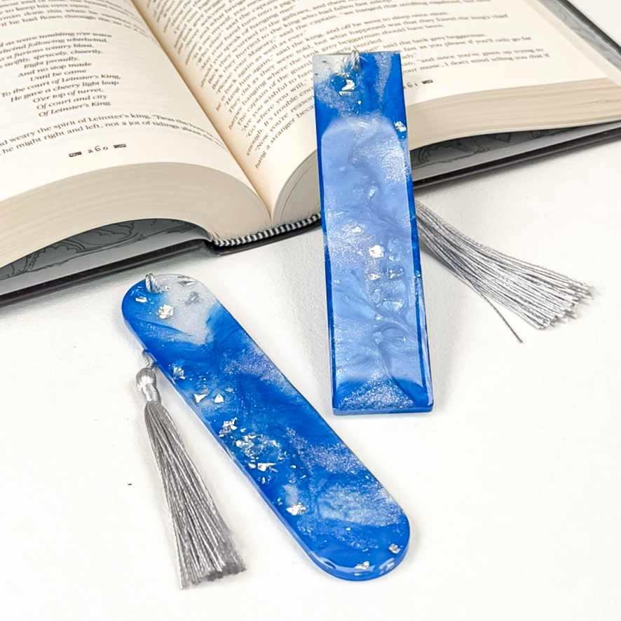 Two blue resin bookmarks with silver tassels, one rectangular and one rounded, displayed on a white surface with an open book in the background.